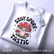 Футболка гриб споретастік білого кольору, зображена зверху, на ній видно принт з грибом та написом 'STAY SPORE-TASTIC', на футболці є етикетка з зеленим шнурком, фон світлий.