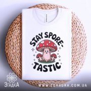 Футболка гриб споретастік білого кольору, складена на плетеній кошику, зображена зверху, фон світлий, принт з грибом та написом 'STAY SPORE-TASTIC' чітко видний.