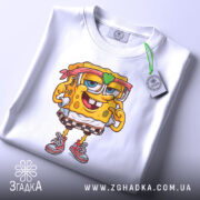 Футболка губка боб білого кольору, складена на столі, з принтом персонажа, на якому видно етикетку з зеленим шнурком, фон білий.