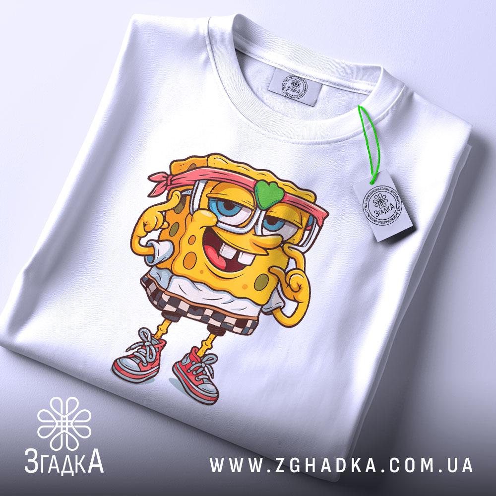 Футболка губка боб білого кольору, складена на столі, з принтом персонажа, на якому видно етикетку з зеленим шнурком, фон білий.