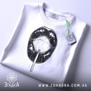 Футболка губи льодяник, біла, складена на поверхні, фон світлий, принт чорного кольору з зображенням губ і льодяника, на комірці є етикетка.