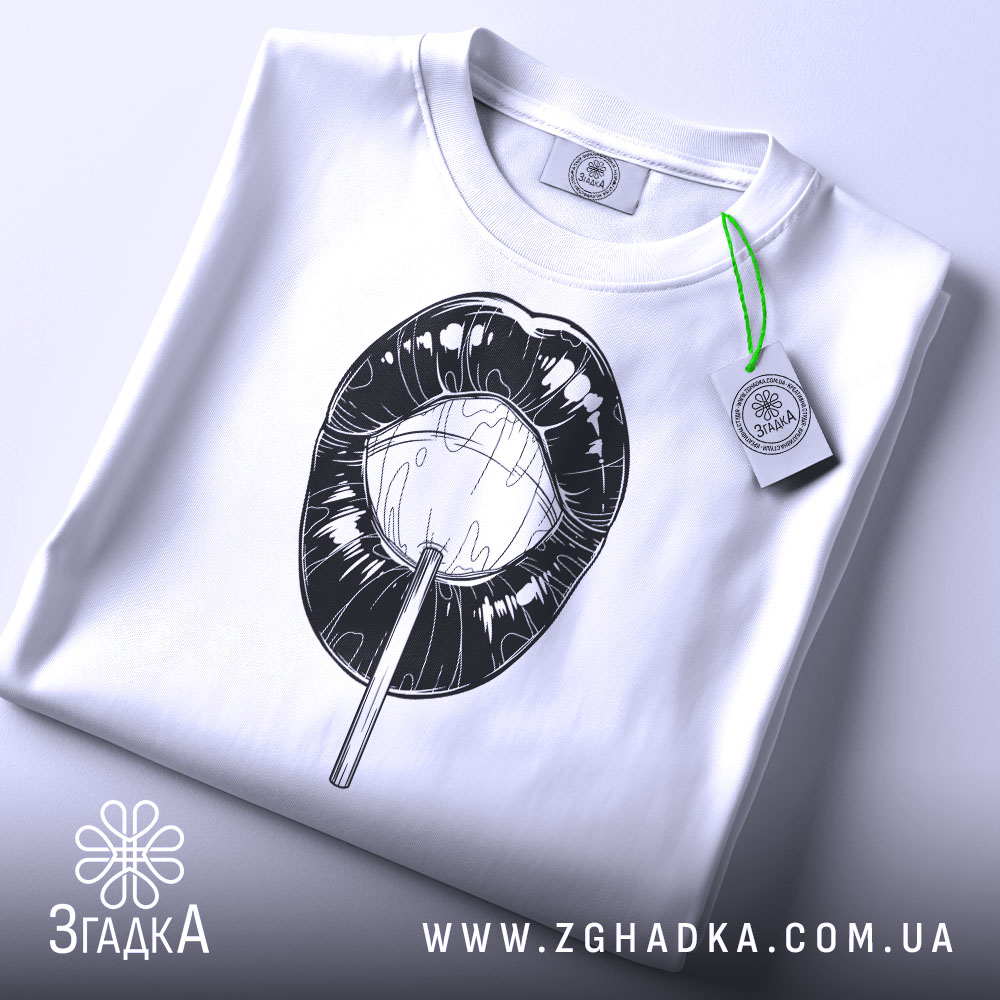 Футболка губи льодяник, біла, складена на поверхні, фон світлий, принт чорного кольору з зображенням губ і льодяника, на комірці є етикетка.