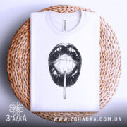 Футболка губи льодяник, біла, складена на кошику з плетеного матеріалу, фон світлий, принт чорного кольору з зображенням губ і льодяника.