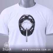 Футболка губи льодяник, біла, складена на поверхні, фон світлий, принт чорного кольору з зображенням губ і льодяника, на комірці є етикетка.