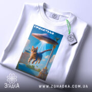 Футболка гумор НЛО, біла, складена на столі, фон нейтральний, принт з котом, що летить під космічним кораблем, яскраві кольори, на футболці є етикетка з зеленим шнурком.