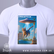 Футболка гумор НЛО, біла, складена на столі, фон нейтральний, принт з котом, що летить під космічним кораблем, яскраві кольори, на футболці є етикетка з зеленим шнурком.