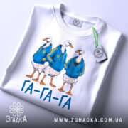 Футболка гуси га га га біла, зображення трьох гусей у синіх худі, лежить на поверхні, принт чіткий, текст "ГА-ГА-ГА" під ними, на футболці є етикетка з зеленим шнурком.