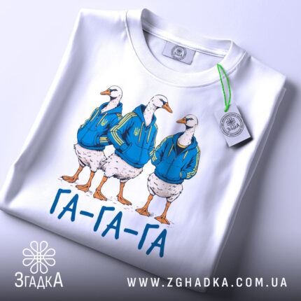 Футболка гуси га га га біла, зображення трьох гусей у синіх худі, лежить на поверхні, принт чіткий, текст "ГА-ГА-ГА" під ними, на футболці є етикетка з зеленим шнурком.