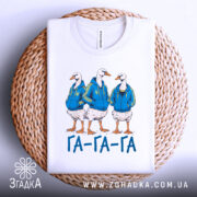 Футболка гуси га га га біла, зображення трьох гусей у синіх худі, лежить на плетеній основі, принт чіткий, текст "ГА-ГА-ГА" під ними, фон світлий.