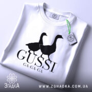Футболка гуси мем, біла, згорнута, верхній ракурс, фон світло-сірий, принт з двома чорними гусями та написом 'GUSSI GA-GA-GA', з етикеткою зеленого кольору.