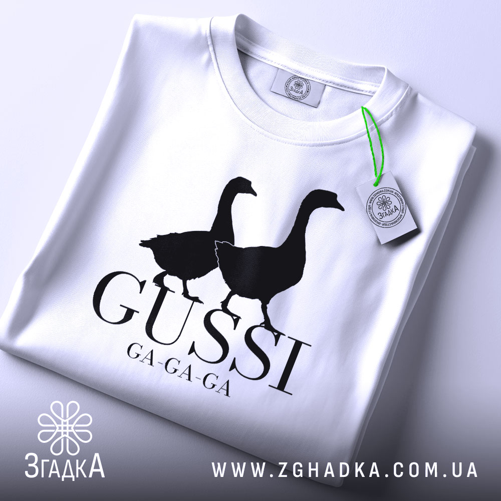 Футболка гуси мем, біла, згорнута, верхній ракурс, фон світло-сірий, принт з двома чорними гусями та написом 'GUSSI GA-GA-GA', з етикеткою зеленого кольору.