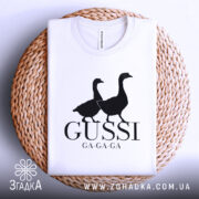 Футболка гуси мем, біла, згорнута, верхній ракурс, фон білий, принт з двома чорними гусями та написом 'GUSSI GA-GA-GA', на плетеній основі.
