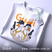 Футболка гуси в кепках білого кольору, складена на поверхні, з видимим етикеткою, фон світло-сірий, принт з двома гусями в кепках та золотими ланцюгами, один гусак тримає пістолет.