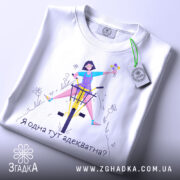 Футболка я адекватна, білого кольору, зображена в складеному вигляді на світлому фоні, принт з дівчиною на велосипеді, яка тримає квіти, текст під малюнком: 'Я одна тут адекватна?', на футболці є етикетка з логотипом.