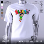 Футболка я алкаш. Біла футболка на манекені, з принтом на грудях, що зображує персонажа в зеленій футболці та жовтих шортах, який стрибає з написом 'ЯЛКАШ' кольоровими літерами. Фон світло-сірий.