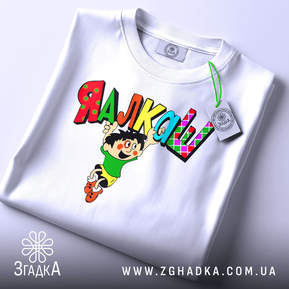 Футболка я алкаш. Біла футболка, складена на столі, з принтом, що зображує персонажа в зеленій футболці та жовтих шортах, який стрибає з написом 'ЯЛКАШ' кольоровими літерами. На футболці видно етикетку.
