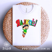 Футболка я алкаш. Біла футболка, складена на плетеному кошику, з принтом, що зображує персонажа в зеленій футболці та жовтих шортах, який стрибає з написом 'ЯЛКАШ' кольоровими літерами. Фон світло-сірий.