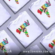 Футболка я алкаш. Біла футболка на манекені, з принтом на грудях, що зображує персонажа в зеленій футболці та жовтих шортах, який стрибає з написом 'ЯЛКАШ' кольоровими літерами. Фон світло-сірий.