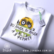 Футболка я фермер. Біла футболка, складена на столі, з видимим принтом, фон світло-сірий. Принт містить жовте сонце, чорний трактор та текст "ВСІ ЛЮДИ, ЯК ЛЮДИ А Я ФЕРМЕР" у чорному та зеленому кольорах, з зеленим ярликом.
