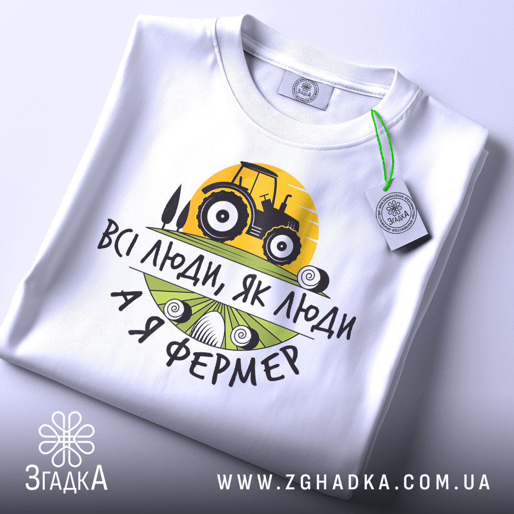 Футболка я фермер. Біла футболка, складена на столі, з видимим принтом, фон світло-сірий. Принт містить жовте сонце, чорний трактор та текст "ВСІ ЛЮДИ, ЯК ЛЮДИ А Я ФЕРМЕР" у чорному та зеленому кольорах, з зеленим ярликом.