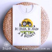 Футболка я фермер. Біла футболка, складена на плетеному кошику, фон світло-сірий. Принт містить жовте сонце, чорний трактор та текст "ВСІ ЛЮДИ, ЯК ЛЮДИ А Я ФЕРМЕР" у чорному та зеленому кольорах.