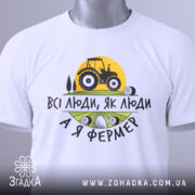 Футболка я фермер. Біла футболка, складена на столі, з видимим принтом, фон світло-сірий. Принт містить жовте сонце, чорний трактор та текст "ВСІ ЛЮДИ, ЯК ЛЮДИ А Я ФЕРМЕР" у чорному та зеленому кольорах, з зеленим ярликом.