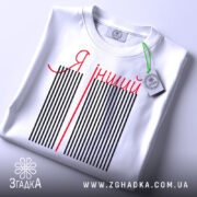 Футболка я інший. Біла футболка, складена на плоскій поверхні, з видимим етикеткою. На футболці чорні вертикальні смуги з червоною написом 'Я інший'. Фон світло-сірий.