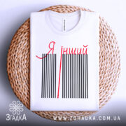 Футболка я інший. Біла футболка, складена на плоскій поверхні з плетеним кошиком на фоні. На футболці чорні вертикальні смуги з червоною написом 'Я інший'. Фон світло-сірий.