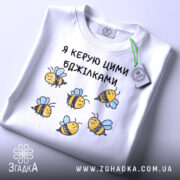 Футболка я керую бджілками, білого кольору, зображена в горизонтальному положенні на світлому фоні, принт з бджілками жовтого кольору з чорними смужками і блакитними крилами, на комірці є етикетка.