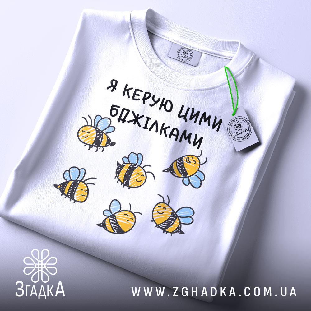 Футболка я керую бджілками, білого кольору, зображена в горизонтальному положенні на світлому фоні, принт з бджілками жовтого кольору з чорними смужками і блакитними крилами, на комірці є етикетка.
