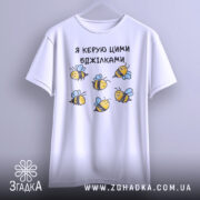 Футболка я керую бджілками, білого кольору, зображена з фронтального ракурсу на світло-сірому фоні, принт з бджілками жовтого кольору з чорними смужками і блакитними крилами.