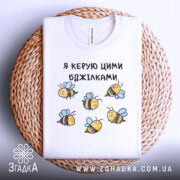 Футболка я керую бджілками, білого кольору, зображена в горизонтальному положенні на плетеній основі, принт з бджілками жовтого кольору з чорними смужками і блакитними крилами.