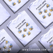 Футболка я керую бджілками, білого кольору, зображена з фронтального ракурсу на манекені, фон світло-сірий, принт з малюнками бджілок жовтого і чорного кольорів з блакитними крилами.