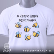 Футболка я керую бджілками, білого кольору, зображена в горизонтальному положенні на світлому фоні, принт з бджілками жовтого кольору з чорними смужками і блакитними крилами, на комірці є етикетка.