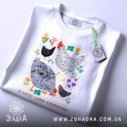 Футболка я керую цими курочками. Біла футболка, зображена зверху, з коротким рукавом. Фон світло-блакитний. Принт з малюнками курей та квітів, яскраві кольори, з текстом внизу. На футболці є етикетка з зеленим шнурком.
