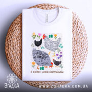 Футболка я керую цими курочками. Біла футболка, зображена зверху на плетеній основі. Фон білий. Принт з малюнками курей та квітів, яскраві кольори, з текстом внизу.