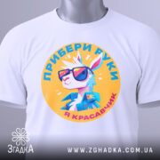 Футболка я красавчик, біла, зображена зверху, на фоні світло-сірого кольору, принт з мультяшним кроликом у сонцезахисних окулярах, текст українською мовою, на етикетці зелена стрічка.