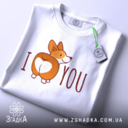 Футболка я люблю білого кольору, зображена в плоскому вигляді, фон світло-сірий, принт з коргі, що показує свою попу, з написом 'I ♥ YOU', на комірці є етикетка.