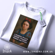 Футболка я людина проста. Біла футболка, складена на поверхні, з видимим етикеткою. Принт з дівчиною в центрі, текст чорного кольору під зображенням. Фон світлий, без зайвих деталей, футболка виглядає охайно.