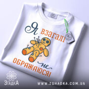 Футболка я не ображаюся, білого кольору, складена на поверхні, зображена з верхнього ракурсу, фон світло-сірий, принт з жовтою лялькою вуду та текстом "Я взагалі не ображаюся!" у помаранчевому та темно-синьому кольорах, на футболці є етикетка.