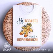 Футболка я не ображаюся, білого кольору, складена на плоскій поверхні, зображена зверху, фон світло-сірий, принт з жовтою лялькою вуду та текстом "Я взагалі не ображаюся!" у помаранчевому та темно-синьому кольорах, футболка лежить на плетеній основі.