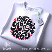 Футболка я обираю себе. Біла футболка, складена на поверхні, з великим принтом чорного кольору і червоними елементами. На футболці є етикетка з зеленим шнурком. Фон світло-сірий, що створює чистий вигляд.