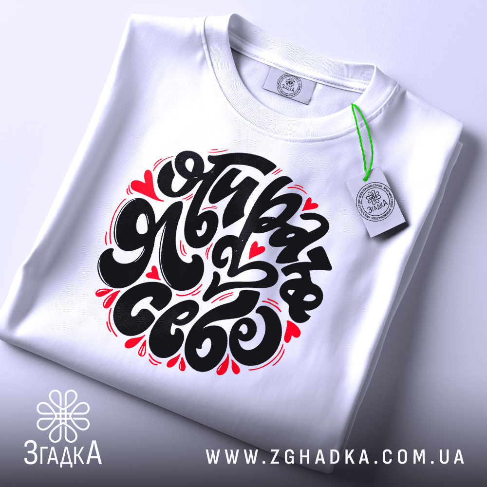 Футболка я обираю себе. Біла футболка, складена на поверхні, з великим принтом чорного кольору і червоними елементами. На футболці є етикетка з зеленим шнурком. Фон світло-сірий, що створює чистий вигляд.