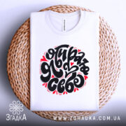 Футболка я обираю себе. Біла футболка, складена на плетеній кошику, з великим принтом чорного кольору і червоними сердечками. Фон світло-сірий, що створює контраст з текстурою кошика.