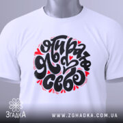 Футболка я обираю себе. Біла футболка, складена на поверхні, з великим принтом чорного кольору і червоними елементами. На футболці є етикетка з зеленим шнурком. Фон світло-сірий, що створює чистий вигляд.