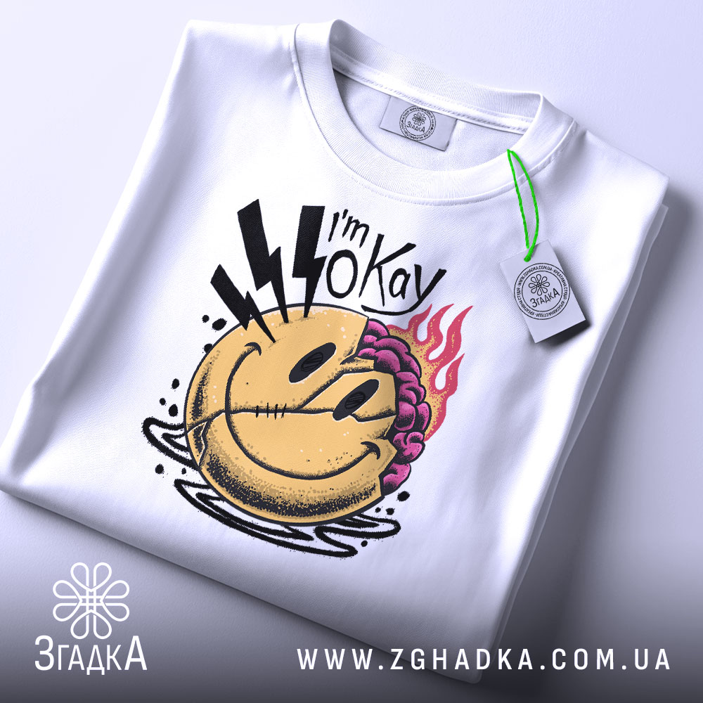 Футболка я окей. Біла футболка, складена на поверхні, з принтом усміхненого обличчя, на якому видно фіолетове волосся та блискавки, з зеленим ярликом, знята з верхнього ракурсу.