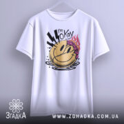 Футболка я окей. Біла футболка з принтом усміхненого обличчя, з фіолетовим волоссям та блискавками, підвішена на вішалці, знята з фронтального ракурсу на світлому фоні.