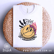 Футболка я окей. Біла футболка, складена на плетеній основі, з принтом усміхненого обличчя з фіолетовим волоссям та блискавками, знята з верхнього ракурсу.