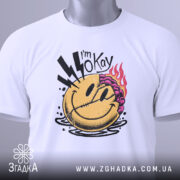 Футболка я окей. Біла футболка, складена на поверхні, з принтом усміхненого обличчя, на якому видно фіолетове волосся та блискавки, з зеленим ярликом, знята з верхнього ракурсу.
