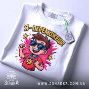 Футболка я переможець, білого кольору, розкладена на поверхні, фон світло-сірий, принт з хлопчиком у сонцезахисних окулярах, який радісно піднімає руку, з написом 'Я - переможець!' та яскравими зірочками, на комірці є етикетка.